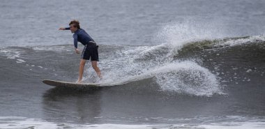 Gilgo Beach, New York, ABD - 30 Ağustos 2023: Genç bir sörfçü sahilde dalgaları yakalarken ustalıkla dengede duruyor.