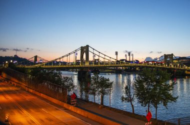 Pittsburgh, Pennsylvania, ABD - 6 Ağustos 2023: Alacakaranlık, nehir boyunca uzanan köprüye sıcak bir parıltı saçarak, canlı şehir ışıklarını ve sakin suyu vurguluyor..