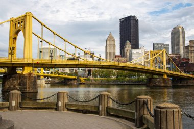 Pittsburgh, Pennsylvania, ABD - 6 Ağustos 2023: Sarı köprü sakin bir nehir boyunca uzanır, bulutlu bir gökyüzünün altında Pittsburgh ufuk çizgisini sergiler..