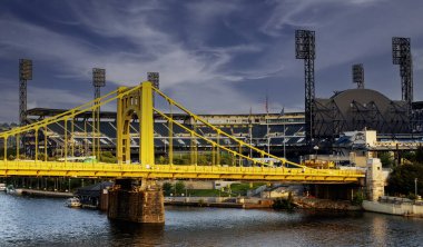 Pittsburgh, Penssilvanya, ABD - 8 Ağustos 2023: Çarpıcı bir sarı köprü nehri geçerek, şehir yaşamını dramatik bir arkaplan ortasındaki bir stadyuma bağlıyor..