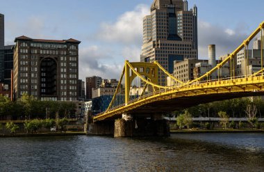 Pittsburgh, Pennsylvania, ABD - 8 Ağustos 2023: Nehrin her iki tarafını birbirine bağlayan canlı sarı bir köprü, açık bir gökyüzü altında hareketli bir şehir manzarası sergiliyor.