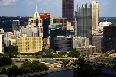 Pittsburgh, Pennsylvania, ABD - 8 Ağustos 2023: Uzun binalar aşağıdaki yemyeşil parklarla nehir boyunca güneş ışığını yansıtarak canlı bir şehir atmosferi yaratıyor.