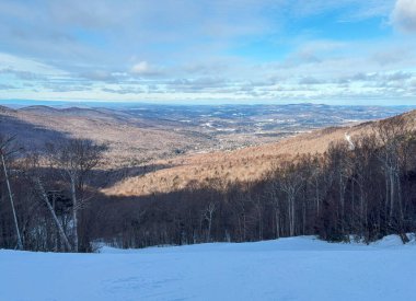 Karla kaplı bir dağ sırası, açık gökyüzünün altındaki vadileri ve uzak tepeleri panoramik bir şekilde gösteriyor..