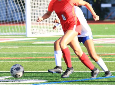 Kırmızı ve beyaz üniformalı iki oyuncu, parlak güneş ışığı altında yeşil bir futbol sahasında bulunma mücadelesi veriyor..