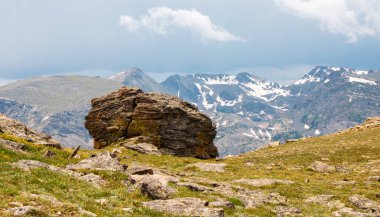 Büyük bir kaya, yüksek dağlar ve yemyeşil ağaçlarla çevrili bir dağlık bölgede belirgin bir şekilde bulunur..