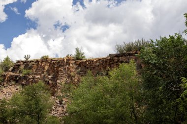 Kayalık uçurum, yeşil ağaçlarla çevrili beyaz bulutlarla kaplı parlak mavi bir gökyüzüne karşı görkemli bir şekilde yükselir..