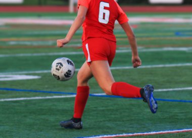 Kırmızı üniformalı genç bir futbolcu, bir maç sırasında sahada ustalıkla topa vuruyor..