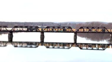 Bir tren, açık bir günde aşağıdaki durgun suda yansıtılan yıpranmış bir köprüde durmadan hareket eder..