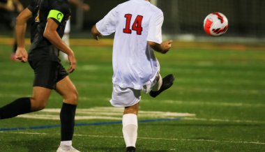 Beyaz üniformalı genç bir sporcu canlı bir sahada futbol topunu tekmeliyor.