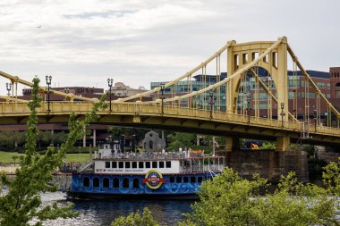 Pittsburgh, Pennsylvania, ABD - 6 Ağustos 2023: Bir tur teknesi şehir binaları arasında parlak sarı bir köprünün altında süzülüyor.