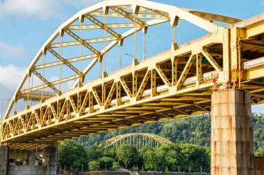 Pittsburgh şehrinde açık mavi gökyüzünün altında büyük sarı bir köprü sakin bir nehri geçer.