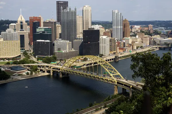 Pittsburgh, Pennsylvania, ABD - 8 Ağustos 2023: Şehir silueti binalar, sarı bir köprü ve sakin bir nehir gösteriyor.