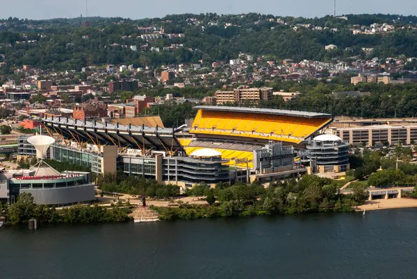 Pittsburgh, Pennsylvania, ABD - 8 Ağustos 2023: Şehir tepeleri ve mavi gökyüzü tarafından çerçevelenmiş nehir boyunca canlı bir spor stadyumu bulunuyor.