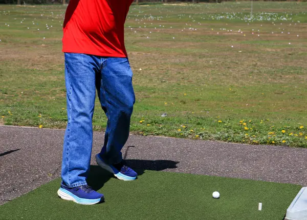 Kırmızı tişörtlü ve kot pantolonlu biri golf sahasında golf topuna vurmaya hazır..