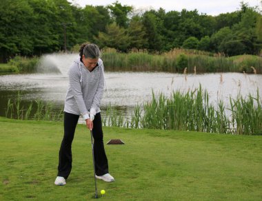 Dişi bir golfçü ağaçlarla çevrili huzurlu bir göletin yakınındaki golf sahasında sallanmaya hazırlanıyor..