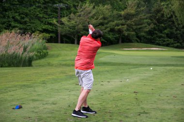 Kırmızı tişörtlü golfçü golf sahasında yeşil çimlerde sallanıyor..