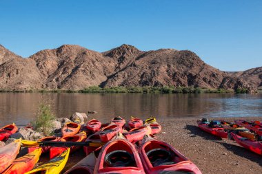 Canlı renklerde kanolar dağlarla çevrili bir nehrin kıyısında dinleniyor..