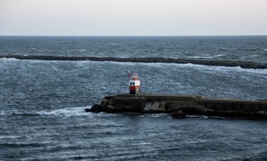 Gün batımının renkleri okyanustan yansıyor. Bir deniz feneri uzaktaki gemileri yönlendiriyor..