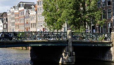 Amsterdam, Hollanda - 5 Ağustos 2025: Amsterdam 'da klasik binalar ve yeşil ağaçlarla çevrili büyüleyici bir kanal köprüsü.