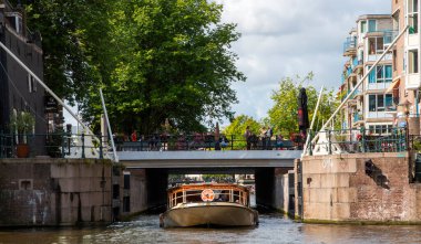 Amsterdam, Hollanda - 5 Ağustos 2025: İnsanlar yukarıdan manzaranın keyfini çıkarırken, bir tur teknesi kanaldaki bir köprünün altında sorunsuzca süzülüyor.