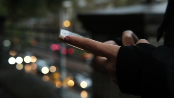 若い女性の手は雨の下で携帯電話にテキストメッセージを送信していると背景にぼやけたトラフィック — ストック動画