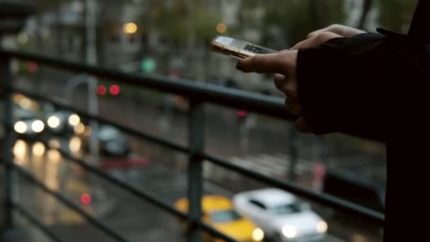 若い女性の手は雨の下で携帯電話にテキストメッセージを送信していると背景にぼやけたトラフィック — ストック動画