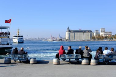 İstanbul, Türkiye - 27 Ekim 2022: Türk halkı ve bazı turistler banklarda oturuyor ve Kadıköy kıyı şeridinde iyi vakit geçiriyorlar. Haydarpaşa Tren İstasyonu ve feribot tekneleri görülebilir..