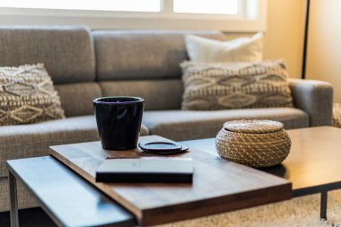 Üzerinde kitap ve kupa olan bir sehpa. Masa oturma odasında, kanepesi ve penceresi var. Sahne rahat ve rahatlatıcı.