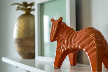 Bir zebra heykeli bir ananas ve çerçeveli bir resmin yanındaki rafta duruyor. Sıcaklık ve samimiyet kavramı, turuncu zebra beyaz rafa renk katıyor.