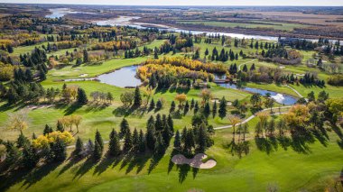 Arka planda ağaçlar ve gölet olan bir golf sahası. Ağaçlar çoğunlukla yeşil ve gölet ağaçlarla çevrili.