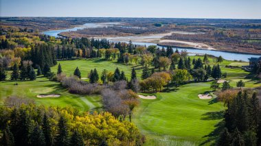 Arka planda nehir olan bir golf sahası. Golf sahası ağaçlarla çevrili ve yeşil çimenli bir alanı var.