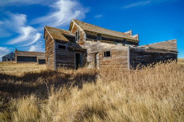 Büyük, eski bir ev uzun otların olduğu bir tarlada duruyor. Ev diğer eski binalarla çevrili ve gökyüzü açık ve mavi. Manzara sakin ve sakin, eski binalarla.