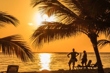 İki kişi sahilde durup gün batımını izliyorlar. Gökyüzü turuncu ve su sakin. Arka plandaki palmiye ağaçları tropikal bir atmosfer sağlar.