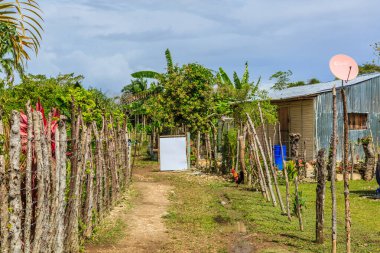 Bir patika, yan tarafında beyaz bir işaret olan bir eve götürür. Evin etrafı çitlerle çevrili ve çatıda uydu anteni var.