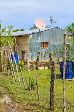 Üzerinde uydu anteni olan küçük bir kulübe. Kulübenin etrafı çitlerle çevrili ve yerde mavi bir fıçı var.