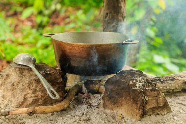 Bir tencere yemek ateşin üzerinde pişiyor, yanında da kaşık var. Sahnenin, basit ve ilkel bir havası var. Basitlik ve doğayla bağlantı hissi uyandırıyor.