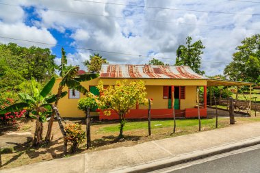 Kırmızı çatılı ve beyaz kapılı sarı bir ev. Ev bir çitle çevrili ve bir verandası var. Ev kaldırımlı bir caddede yer alıyor.
