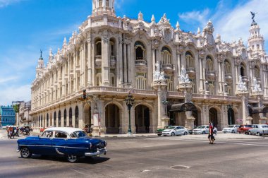 A blue car is driving down a street in front of a large building. The street is busy with cars and people walking. The scene has a nostalgic and classic feel, with the old car clipart