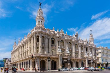 Üstünde bir sürü pencere ve heykel olan büyük bir bina. Bina beyaz ve çok fazla detayı var. Gökyüzü mavi ve birkaç bulut var.