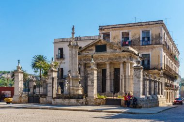 Beyaz yüzlü ve siyah çitli büyük bir bina. Binanın önünde büyük bir kemer yolu ve bir heykel var. Sokaklar boş ve etrafta hiç insan yok.