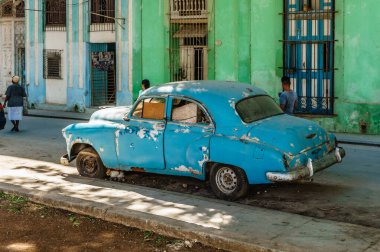 Mavi bir araba bir binanın önündeki caddede park halinde. Araba eski ve üzerinde çok paslı bir şey var. Ortalık sakin ve sakin, etrafta kimse yok.