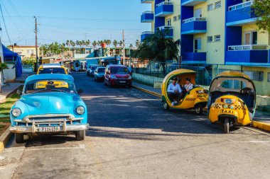 Yolun kenarına park edilmiş mavi bir araba sarı bir taksinin yanında. Taksi açık ve içeride iki adam oturuyor. Sahne, eski ve yeni arabaların karışımı olan bir şehirde geçiyor.