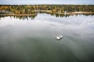 Bir ormanın önündeki suda bir tekne var. Gökyüzü bulutlu ve su sakin.