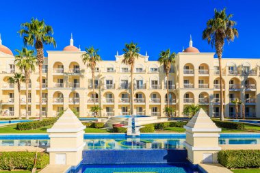 Önünde bir havuz ve palmiye ağaçları olan büyük bir bina. Binanın bir sürü penceresi ve balkonu var.