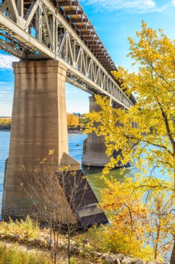 Bir köprü aşağıdaki su manzaralı bir nehri kapsar. Köprü beton ve çelikten yapılmıştır. Ağaçlardaki yapraklar sarı, bu da sonbahar olduğunu gösterir.