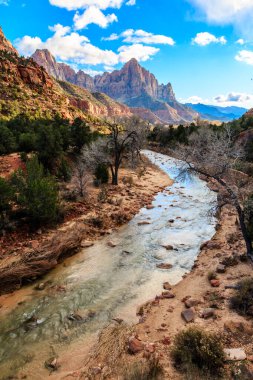 Arka planda dağlar olan bir nehir bir kanyonun içinden akar. Su berrak ve sakin, nehir kenarındaki ağaçlar da çıplak. Huzurlu ve huzurlu bir sahne.