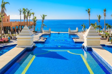 Arka planda mavi okyanus olan güzel bir havuz. Havuz palmiye ağaçlarıyla çevrili ve beyaz bir çatısı var.