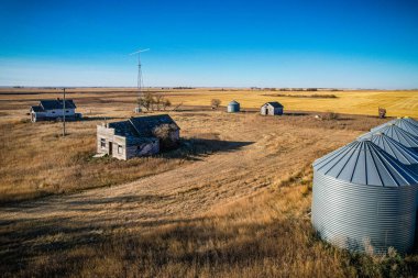 Birkaç eski evi ve birkaç silosu olan bir kırsal bölge. Evler eski ve terk edilmiş. Silolar da boş. Gökyüzü mavi ve çimenler kuru.