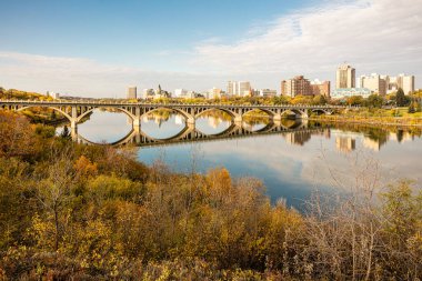 Bir köprü, arka planda bir şehir olan bir nehri kapsar. Köprü ağaçlarla çevrili ve su sakin.