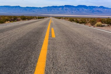 Ortada sarı bir çizgi olan uzun, boş bir yol. Yol dağlarla çevrili ve gökyüzü mavi.